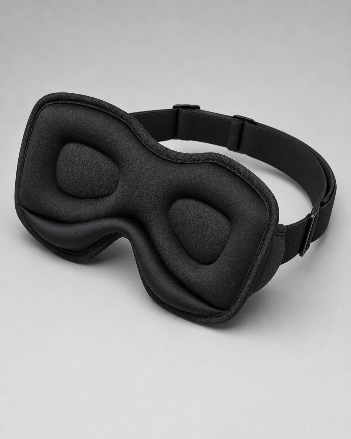 A black padded sleep mask with an adjustable strap, photographed on a clean light gray studio background.