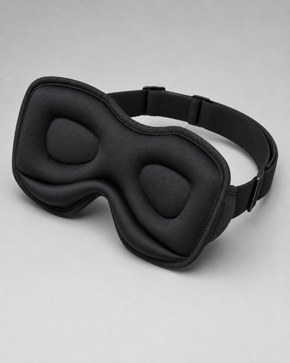 A black padded sleep mask with an adjustable strap, photographed on a clean light gray studio background.